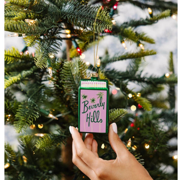 Hand holding a decorative ornament with 'Beverly Hills' text against a Christmas tree background