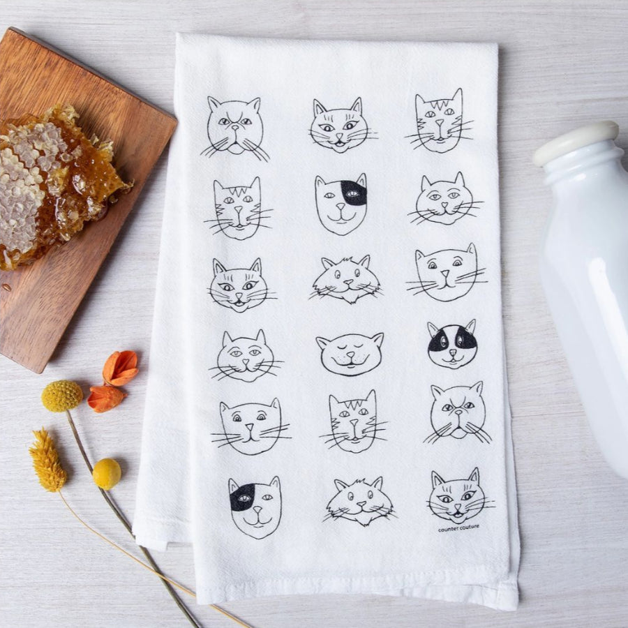 White towel with black cat illustrations on a wooden surface with a bottle and flowers.