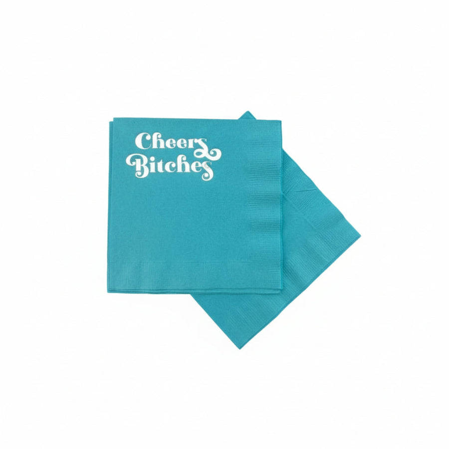 Turquoise napkin with 'Cheers Bitches' text on a textured white background