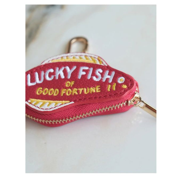 A small red zippered pouch shaped like a fish head, embroidered with 'LUCKY FISH OF GOOD FORTUNE 11' in white and yellow, shown on a light marble surface.