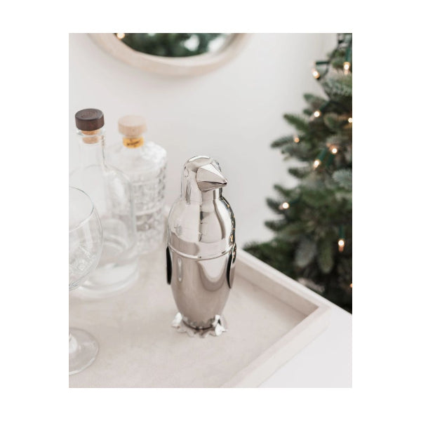 A polished silver penguin-shaped cocktail shaker resting on a light beige tray, with clear glass bottles and a softly blurred Christmas tree in the background.