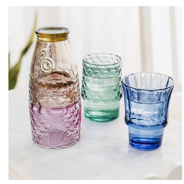 Three textured glass drinking vessels on a white surface: a tall bottle with a gold-toned rim in translucent pink-tinted glass, a small green-tinted tumbler, and a blue-tinted tumbler, against a soft white curtain background.