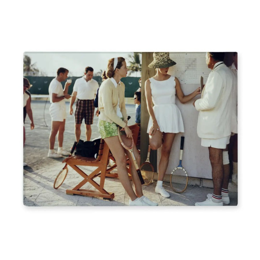 Vintage scene of people on a tennis court with one woman in a white dress holding a tennis racket.