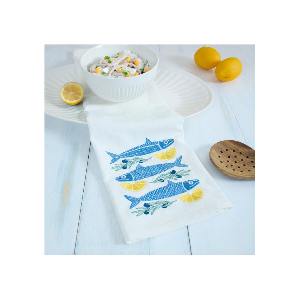 A white kitchen towel with embroidered blue fish and yellow lemon slices, laid on a light blue wooden table next to a white plate with salad, lemons, and a wooden spoon.